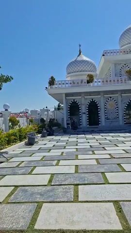 A man stands outdoors in front of a mosque with two domes. He is wearing a dark jacket and light-colored pants and is touching his hair. The mosque has "MASJID HARTON" written on its facade. In front of the mosque is a paved courtyard with square stone tiles and patches of grass. There are trees and other vegetation in the background, as well as cars and a pole with lights. The sky is clear and blue.
