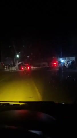 The video displays a night-time road scene viewed from the interior of a vehicle, looking through the windshield. The vehicle's dashboard, with red illuminated indicators, is visible at the bottom of the frame. Bright yellow headlights from the recording vehicle illuminate the asphalt road ahead, revealing a yellow center line. In the distance, multiple vehicles are present, with two vehicles prominently displaying flashing red lights, possibly indicating hazard lights or emergency vehicles. Other ambient streetlights and vehicle lights are scattered throughout the dark environment.