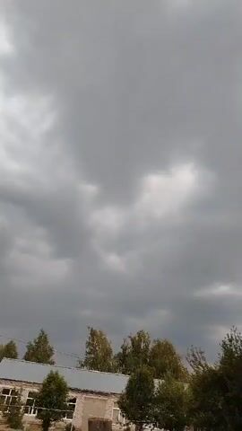 The video captures a cloudy sky with trees and a building in the foreground. The clouds are dark and appear to be moving, suggesting a possibly stormy or overcast day. The trees are green and leafy, indicating it is likely a warmer season. A building, possibly a school or community center, is visible with a brick facade. The overall scene is mundane and depicts an outdoor environment.