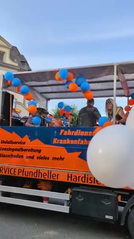 A mobile stage, decorated with orange and blue balloons, is moving down a street. On the stage, a person with blonde hair and sunglasses is playing a guitar. Another person, wearing a white t-shirt, is standing and gesturing with their hand. A man in an orange t-shirt and shorts walks past the front of the truck. Two people, one wearing a pink angel wing costume and another with white angel wings, walk behind the truck. The truck has text that appears to be in German. Several large white balloons are attached to the front of the truck. The sky is clear and blue, and the sun is setting.