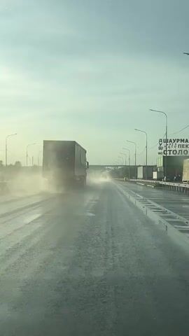 The video shows a view from inside a vehicle driving on a wet highway. A large truck is in front, kicking up a spray of water as it drives. The sky is overcast and appears to be late in the day or early morning due to the lighting. Streetlights and roadside barriers are visible along the highway. There is also a billboard in the distance.