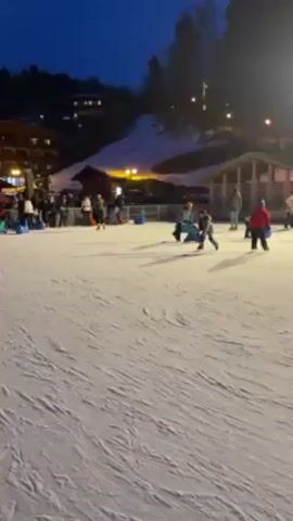 The video shows an outdoor ice rink at dusk, surrounded by people. Some people are ice skating, and others are watching from the sidelines. The rink is surrounded by barriers and the ground is covered in snow. There are buildings and trees visible in the background. The sky is a deep blue. One child falls while skating near the edge of the rink.