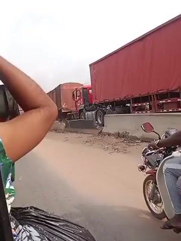 The video shows several large trucks on a road with barriers. Other vehicles, including a silver car and a smaller truck with a colorful body, are also visible. A person's arm appears in the foreground of the video. The environment appears to be somewhat dusty or sandy, and the sky is overcast.