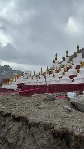 The video shows a group of small, white Buddhist stupas with golden spires. They are built on a red base and arranged in rows on a hillside. A red ribbon runs in front of the base. The ground in front of the stupas is rocky and uneven. Behind the stupas, a steep, rocky mountain rises towards a cloudy sky. There is some audible conversation, but it is unintelligible.