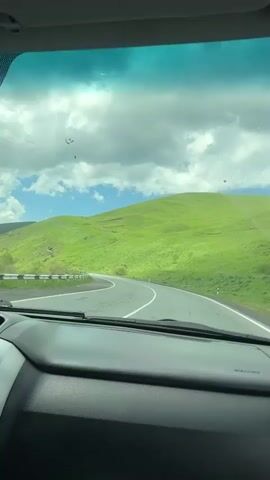 This video shows a point-of-view shot from inside a car looking out the windshield. The car is driving on a paved road that curves to the left. The road is surrounded by lush green hills under a partly cloudy sky. Power lines are visible against the sky. The dashboard of the car is partially visible in the foreground.