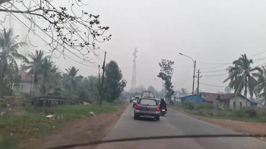 The video shows a view from inside a vehicle driving on a two-lane road. The sky is overcast and hazy, suggesting poor air quality or fog. The road is bordered by vegetation, including palm trees and other trees, and small houses. Utility poles with power lines are visible along the roadside. Several vehicles are on the road, including cars and motorcycles. A tall transmission tower is visible in the distance. The camera appears to be mounted on the dashboard or windshield of the vehicle. The overall impression is of a rural or suburban area with ongoing traffic.