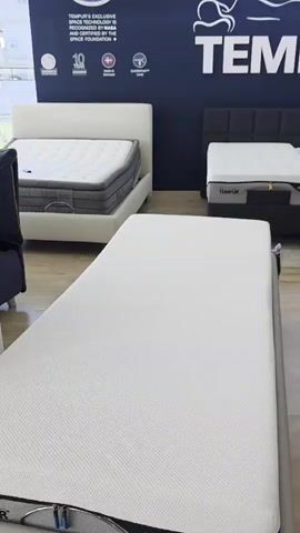 The video shows a retail environment, likely a mattress store. The dominant brand is Tempur, with their logo and branding visible on the dark blue wall. Several mattresses are displayed on beds. In the foreground, a plain white mattress is laid out horizontally. To the left, a white mattress is on a bed frame. In the background, two mattresses are side-by-side on dark bases, with one appearing to be adjustable. Further to the right, a larger bed with a textured headboard and a patterned mattress is visible. The lighting is bright, illuminating the showroom floor, which appears to be made of wood laminate. A window with glass blocks is present near the ceiling. An image of an astronaut is visible on the wall in the background. There are no people visible in the video.