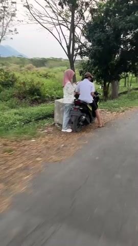 A man and a woman are standing next to a motorcycle on the side of a rural road. The man is sitting on the motorcycle, and the woman is standing beside him. Both are looking away from the camera. The motorcycle is black and has a license plate. The woman is wearing a pink hijab and a white shirt. The man is wearing a white t-shirt and a baseball cap. The road is paved, and there is a ditch with some water on the side. There are trees and bushes in the background, and a mountain in the distance. The sky is overcast.