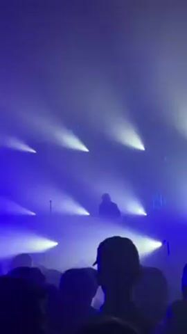 The video shows a concert crowd, mostly blurred and in silhouette, facing a stage illuminated with bright white spotlights shining through a haze of fog. On stage, an individual stands behind what appears to be a DJ setup, also silhouetted against the lights. The overall color scheme is blue, with the spotlights creating a stark contrast against the darker background.