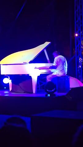 The video captures a live piano performance on a stage. The central focus is a man playing a white grand piano under vibrant stage lighting, primarily in shades of blue and purple. The lighting casts dramatic shadows and highlights on the piano and the performer, creating a theatrical atmosphere. The performer is a man dressed in a light-colored shirt and pants, seated on a dark stool. He is actively playing the piano, with his hands on the keys.

The setting appears to be an outdoor venue at night, given the dark backdrop and the presence of stage lighting. In the background, a tall metal structure, possibly part of the stage rigging or a nearby building, is visible. The overall mood is one of focused musical performance. The audience is partially visible in the foreground, represented by silhouettes, indicating that this is a public event.

The timestamp "00:00" suggests the beginning of the video clip. There is no visible text within the frame. The setting is identified as Cornellà de Llobregat, Spain. The event is a piano concert.