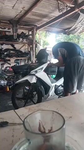 A person wearing a dark blue t-shirt and grey shorts is working on a white motorcycle in a dimly lit workshop. The person is leaning over the front of the motorcycle, appearing to be adjusting something near the handlebars. In the foreground, out of focus, is a glass with a dark liquid. Shelves filled with motorcycle parts are visible in the background. The ceiling is made of corrugated metal.