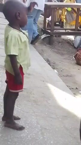 The video shows a busy street scene, possibly in an African country.  In the first shot, a small child wearing a green shirt and red shorts stands barefoot on the pavement, looking off to the side.  The camera then pans to a group of people, mostly women in traditional Muslim dress, sitting or standing near a yellow motorized vehicle (possibly a tuk-tuk). A young man is seated, preparing what appears to be a snack with a small spoon and a white stick or utensil. Several women are around him, some carrying small cups, possibly waiting to receive food. A woman in a purple robe has a baby in a carrier on her back, with the baby's foot visible.