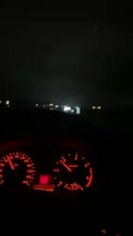 A car travels on a road at night. The dashboard is lit with red lights and indicates the car is traveling at around 100 km/h. The windshield wipers are on. The road ahead is wet and illuminated by headlights from other cars.