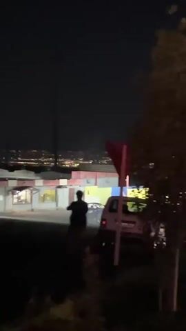 At night, a silhouetted figure is seen walking on a hill with a cityscape in the background. The figure is near a sign and some trees. A building with a red roof and a couple of parked cars are also in the background. A beam of green light is briefly seen in the sky.