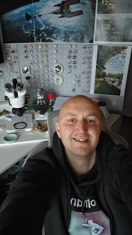 A bald man is sitting in a chair, smiling at the camera. He is wearing a black hooded sweatshirt. Behind him is a desk with two microscopes, jars, and other scientific items. A bulletin board with various pins and images is visible on the wall behind the desk, along with framed images of space and nature. There is also a window with a blurry view of the outside. The camera appears to be held at arm's length in a selfie position.