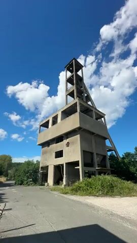The video captures an abandoned concrete structure resembling a mine headframe. The tower-like structure is a stark silhouette against a bright blue sky dotted with fluffy white clouds. The concrete is weathered and shows signs of decay, with empty window openings and structural supports visible. Overgrown green vegetation, including bushes and trees, surrounds the base of the structure and encroaches on a paved road leading towards it. The overall scene suggests a derelict industrial site in a rural or semi-rural setting under a clear, sunny day. There are no people or discernible actions taking place.