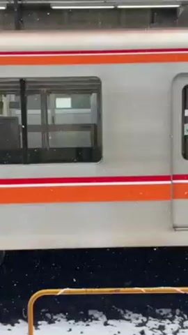 The video shows a train car at a train station in a snowy environment. The train car is primarily light gray with an orange and red stripe running horizontally along the side. The train car's door opens and then closes. The number 2406 is printed on the side of the train car. Some snow is accumulating on the ground. An audible announcement is made, though the specific words are not clear.