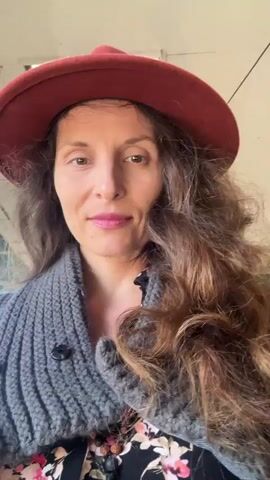 The video is a close-up shot of a woman with long, wavy brown hair and a red hat. She is wearing a gray knitted cardigan over a floral top. Her eyes are closed at the beginning of the video, then she opens them and smiles. The background appears to be an indoor space with plants and a disco ball visible. The lighting is bright, and the overall atmosphere is relaxed and cheerful.