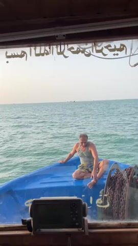 A man sits on the bow of a blue boat with a chain on the right side. He is wearing a tank top and shorts. The sea is visible in the background, with a hazy sky. There is text in Arabic script written on the window above the man. A black electronic device is in the foreground, mounted on the boat.