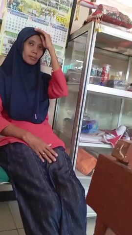 A woman wearing a blue hijab and a red shirt with dark patterned pants sits on a green cushion in a room. A calendar hangs on the wall behind her. To her right is a glass display cabinet filled with various items. The woman appears to be looking at the camera and touching her hair.
