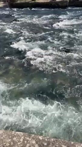 The video depicts a rapidly flowing river or stream. The water is turbulent, with numerous whitecaps and frothy areas. The river appears to be flowing over rocks or a shallow riverbed. The water is generally dark with hints of green or blue.  The edges of the river are lined with a stone or concrete embankment. The overall impression is of a powerful current in a natural or slightly modified watercourse.