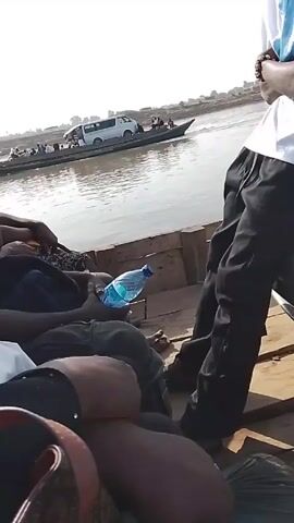 The video shows a group of people traveling on a boat, possibly a ferry. The camera is positioned low, focusing on people sitting or lying down. A plastic water bottle rests near one person. In the background, another boat, loaded with people and a white van, is visible on the water. The environment appears to be a river or lake on a sunny day. The foreground boat is made of wood and seems simple in construction.