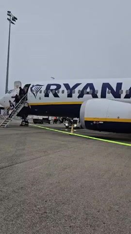 The video shows a Ryanair airplane parked on an tarmac. A set of stairs is connected to the aircraft's entrance, and several people are seen on the stairs and near the plane. A baggage cart is also visible. The ground is paved, and the sky is overcast. A bright green line is visible on the tarmac in the foreground.