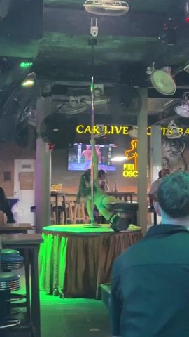 The video shows a woman performing a pole dance on a small stage in a bar. She is wearing a blue bikini, black boots, and black stockings. The stage is covered with a gold-colored tablecloth. A security camera is mounted on the ceiling. In the background, people sit at tables, and a television screen shows a wrestling match. The bar has a neon sign that reads “CAR LIVE SPORTS BAR.” The lighting is dim, with colorful lights accentuating the stage.