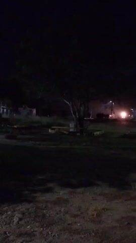 A dark outdoor scene at night. In the center is a large tree with a dark canopy. In the background, there are bright headlights from what appears to be a vehicle approaching or passing by. Smoke or dust is visible in the air around the headlights. To the left of the tree, there are some low, possibly yellow, structures, and to the right, a street light. The foreground is dark, with visible shadows on the ground.