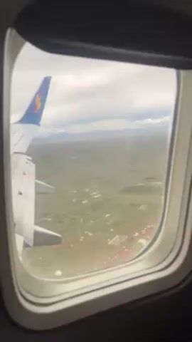 A view through an airplane window shows the ground far below. A wing of the plane is visible in the lower left. The ground is mostly green with many small buildings with red roofs, and a few yellow structures. A mountain range is barely visible in the distance. The sky is cloudy.