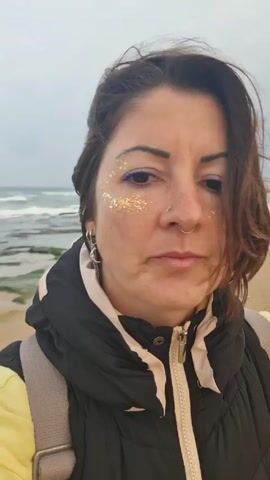 A person with shoulder-length brown hair is taking a selfie video on a beach. The person has fair skin, with glitter makeup around their eyes, a nose ring, and earrings. The person is wearing a black vest with a yellow lining and a backpack. The hair is being blown by the wind, which is strong enough to make the person squint. The ocean is visible in the background with gentle waves, and the sky is overcast. The sand is visible in the foreground.