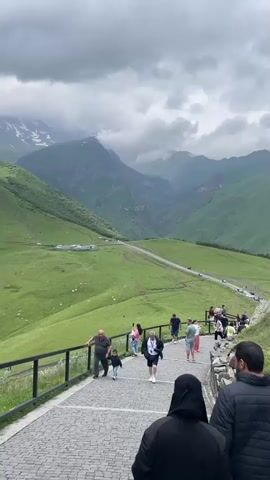 The video shows a group of tourists walking up a paved, upward-sloping path lined with a wooden railing. Several people, including adults and children, are visible on the path. In the background, there are lush green, rolling mountains under a cloudy sky. The path leads towards a broader area where more people are gathered and a road with vehicles can be seen winding through the landscape. The scene is set in a mountainous, natural environment, likely a tourist attraction.