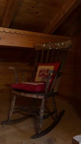 This media file shows a still shot of an antique wooden rocking chair. The chair is adorned with a red cushion and a smaller decorative pillow featuring a red and gold pattern with white reindeer silhouettes. The chair is positioned in a room with wooden walls and ceiling, suggestive of a rustic cabin or attic space. The lighting is dim, creating a warm and cozy atmosphere. The rocking chair's curved runners are visible on the wooden floor, indicating it's ready for use. The overall scene evokes a sense of peace and tranquility. The image is likely captured indoors, given the confined space and wooden construction. There is no visible text in the media file.