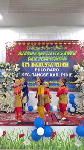 A group of five young children are performing a dance on a stage. The children are wearing traditional Indonesian attire. The stage is decorated with a banner that reads "Selamat dan Sukses Ajang Kreativitas Anak dan Perpisahan RA DARUSSA'ADAH PULO BARO KEC. TANGSE KAB. PIDIE". There are also balloons and floral decorations on the stage and surrounding areas. The children are moving in unison and appear to be enjoying themselves. The overall atmosphere is festive and celebratory.