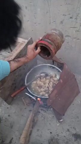 A person with dark curly hair is shown from behind, wearing a light blue shirt. They are pouring liquid from a round, red and brown container into a metal pot. The pot contains pieces of food being cooked over an open fire. The fire is fueled by wood and is situated on a dirt or concrete ground. A makeshift windbreak, made of a brown material, surrounds the cooking area. Another person's legs are visible at the bottom of the frame, wearing what appear to be light-colored shoes or sandals. The environment appears to be outdoors, with a textured wall in the background. The camera is positioned at a slightly elevated angle looking down on the scene.