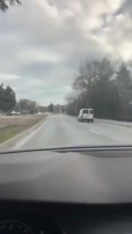 The video shows a view from inside a car driving on a paved road. A white van is driving in the same direction ahead of the car. On either side of the road is grassy areas with bare trees and bushes. Further in the distance are buildings. The sky is cloudy.