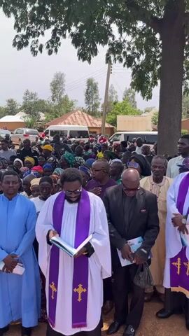 A group of people is gathered outdoors for what appears to be a funeral service. Several clergy members, wearing robes and stoles, are standing at the front, one of whom is reading from a book. A crowd of people is gathered behind them. Some of the people are holding service programs. A casket is visible at the front, adorned with flowers. The setting appears to be a rural area, with houses and vehicles visible in the background. The sky is overcast.