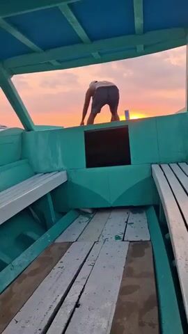A person in shorts and a light-colored shirt is bending over on the deck of a turquoise boat. The boat has wooden benches on either side of a central walkway. The person appears to be engaged in some task. Behind the person, the sun is setting over the water, casting a warm orange and pink glow across the sky. Other boats are visible in the distance. The camera is positioned inside the boat, looking out towards the sunset.