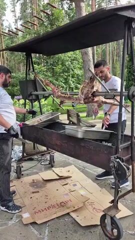 Two men are preparing to roast a whole animal, possibly a goat or lamb, on a spit over a large charcoal grill. The animal is already on the spit and is positioned above the grill. The man on the left, wearing a white t-shirt and striped pants, is holding the spit with his gloved hands. The man on the right, wearing a white t-shirt and black pants, is holding a utensil and appears to be tending to the grill or the animal. A black canopy covers the spit and grill setup. The scene is outdoors, with trees and foliage in the background, suggesting a park or wooded area. Several pieces of cardboard are on the ground around the grill, one with some text that is partially visible. There are metal trays and other cooking equipment on or near the grill. The overall impression is of an outdoor event or catering setup focused on roasting a large piece of meat.