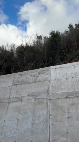 The video shows a concrete retaining wall with a forested hill above it, filmed from a moving vehicle. The concrete wall is textured and grey, with visible seams and bolt holes. Above the wall, there is a slope of dirt and rocks leading up to a line of trees. The trees are mostly bare, with some green foliage mixed in. The sky is visible above the trees, with white clouds against a blue background. The camera is moving along the wall, capturing the details of the concrete and the natural landscape above it.