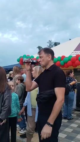 A man in a black polo shirt drinks from a bottle as a woman stands beside him. In the background, a crowd gathers near a building adorned with red and green balloons.