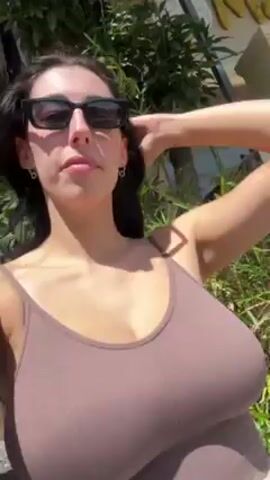The video features a woman with dark hair and sunglasses, wearing a brown tank top and a white skirt. She is standing outside, initially near greenery, then on a brick-paved area. The woman smiles and waves at the camera. The video is taken from a close-up, selfie perspective.