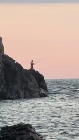 A video shows a man fishing from atop a rocky cliff in a body of water, possibly the sea. The man, wearing a light-colored shirt and dark shorts, stands on the cliff, holding a fishing rod. The sky behind him is colored a pastel pink, suggesting either sunrise or sunset. The sea has gentle waves. The cliffs are dark and appear rugged. The camera remains steady and at a distance throughout the clip.