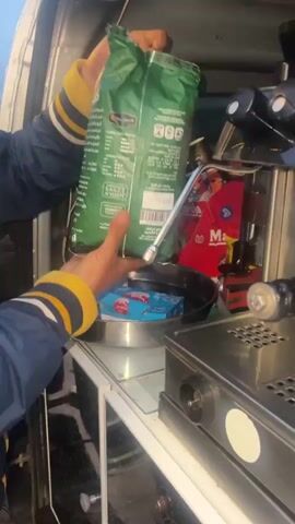Here is a description of the video:
A person standing in a vehicle prepares to make coffee using a professional espresso machine. The person, wearing a blue jacket with yellow and white stripes on the sleeve, holds two green bags, presumably containing coffee beans. The bags are placed on a countertop within the vehicle. Various coffee-making supplies, including milk cartons and boxes of snacks, are visible in the background. The espresso machine is stainless steel, with black knobs and levers, sitting atop the counter. The person's actions are centered around preparing the espresso machine, indicating an intention to brew coffee. The scene is brightly lit, showing the inside of the vehicle, which appears to be set up as a mobile coffee station.
