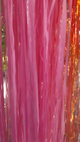 The video captures a close-up view of shiny, metallic streamers in shades of pink, red, and gold. The streamers appear to be hanging, and the camera moves through them, creating a blurred effect. The background is out of focus, but suggests an outdoor environment with greenery and sunlight. The movement of the camera creates a sense of depth and visual texture. There are no people or discernible objects in focus.