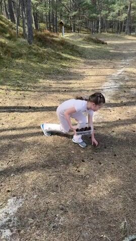 A young girl is picking up pine cones in a forest. She is wearing a light pink t-shirt and pants, and blue sneakers. She has a braided hairstyle. She is carrying a clear container full of pine cones. She picks up pine cones off of the ground. The ground is covered in pine needles and cones. There are tall pine trees surrounding the area.