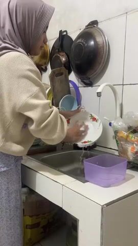 A young woman wearing a light purple hijab and a cream-colored sweater is washing dishes in a sink. She is facing away from the camera, towards the sink. She is holding a white plate with a red floral design and scrubbing it with a sponge. Water is running from a white faucet into the sink. To the right of the sink is a purple plastic container. The walls behind her are tiled in white and there are several pots and pans hanging on the wall. The counter top appears to be made of white tiles. A cardboard box is visible below the counter.