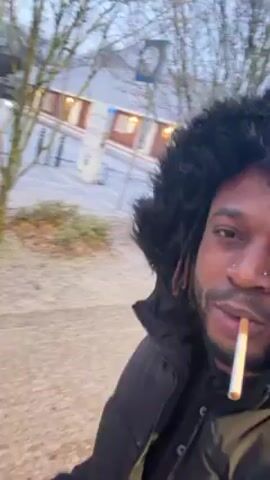 A person with dark hair and a black jacket is holding a cigarette in their mouth and looking at the camera. The person is standing on a snow-covered ground. Behind the person, there are trees, a building, and cars covered in snow. The sky is light blue.