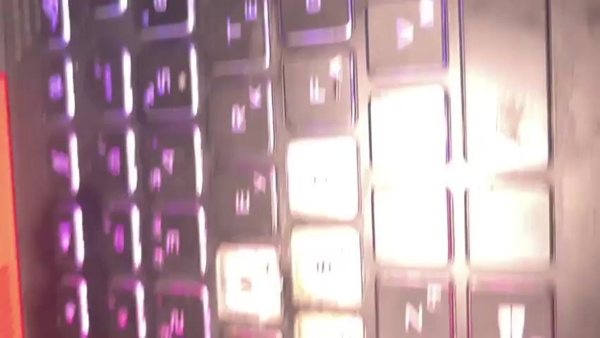 The video displays a close-up, rotating view of a black laptop keyboard. The keyboard features illuminated keys with a mix of purple, blue, and green lighting, suggesting customizable RGB backlighting. The keys show some wear and dust. Visible keys include letters, numbers, function keys like PRTSC, and navigation keys such as arrow keys, END, PGUP, and PGDN. On the left side of the keyboard, a red power indicator light and a USB port are visible. The overall impression is of a gaming or high-performance laptop.