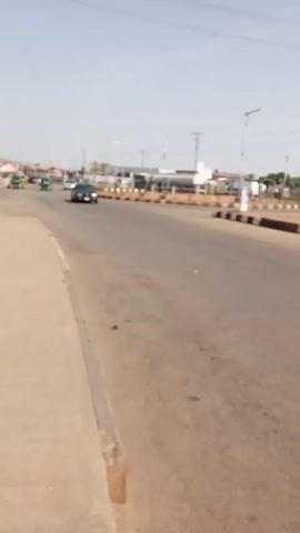 The video shows a street scene on a sunny day. The road is wide with a concrete sidewalk on the left. A black sedan drives by, followed by a green auto rickshaw. Several light poles line the road. In the distance, buildings and vegetation are visible. The sky is pale blue.